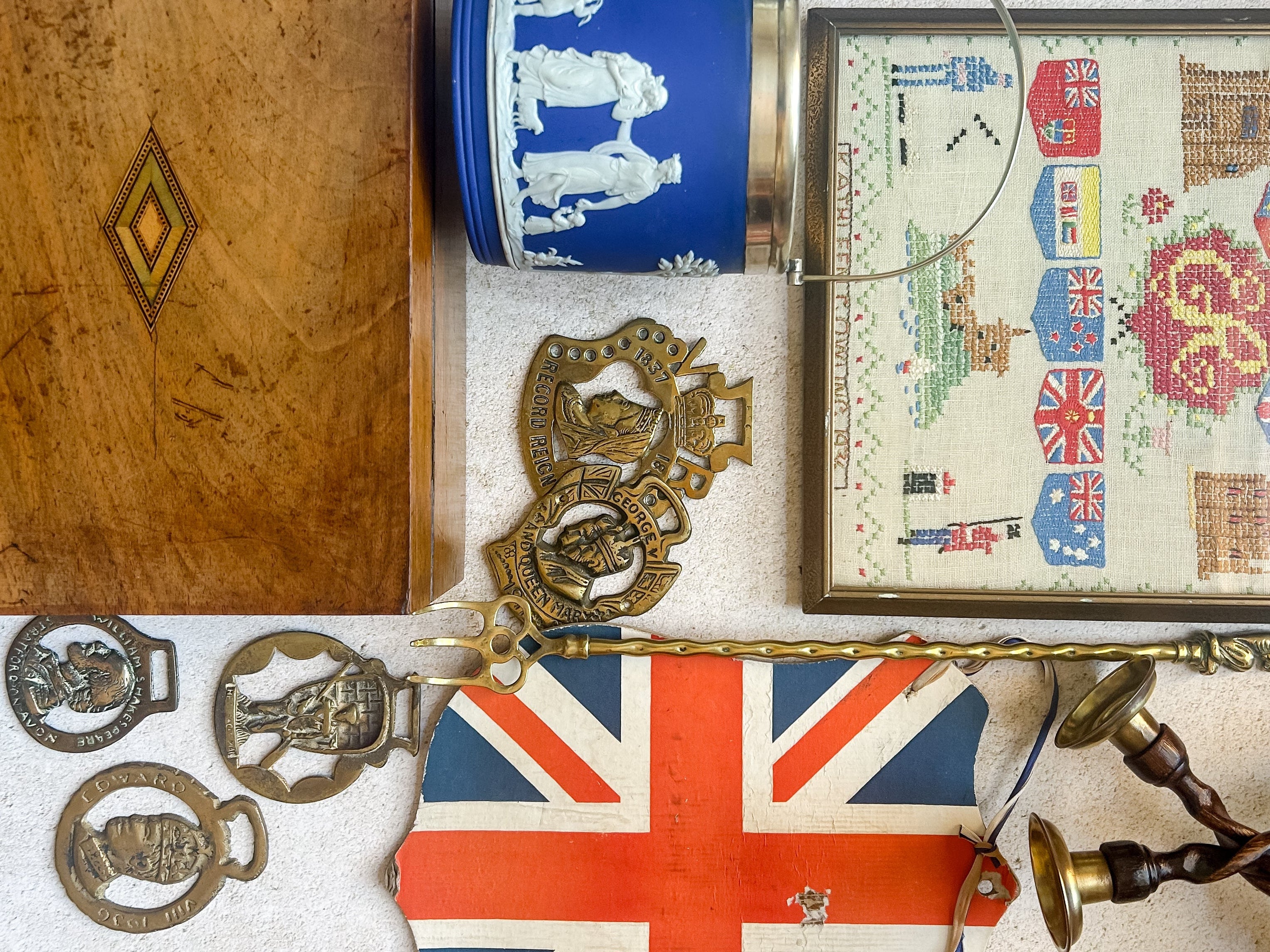 A styled vintage display featuring a framed cross-stitch sampler with British flags, a blue and white porcelain canister, a wooden chest, and several antique bronze military medals against a textured wall.