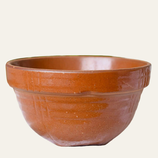 A vintage brown ceramic crock bowl with a geometric pattern and a squared off base, displayed on a white surface with a green plant in the background.