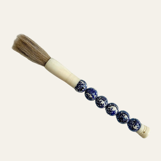 A traditional Chinese calligraphy brush with natural brown bristles, a cream-colored wooden handle, and a string of blue and white patterned ceramic beads, displayed diagonally on a light gray textured background.