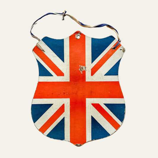 A vintage shield-shaped wooden wall sign featuring the Union Jack flag, with a hanging loop at the top, shown against a textured white wall.