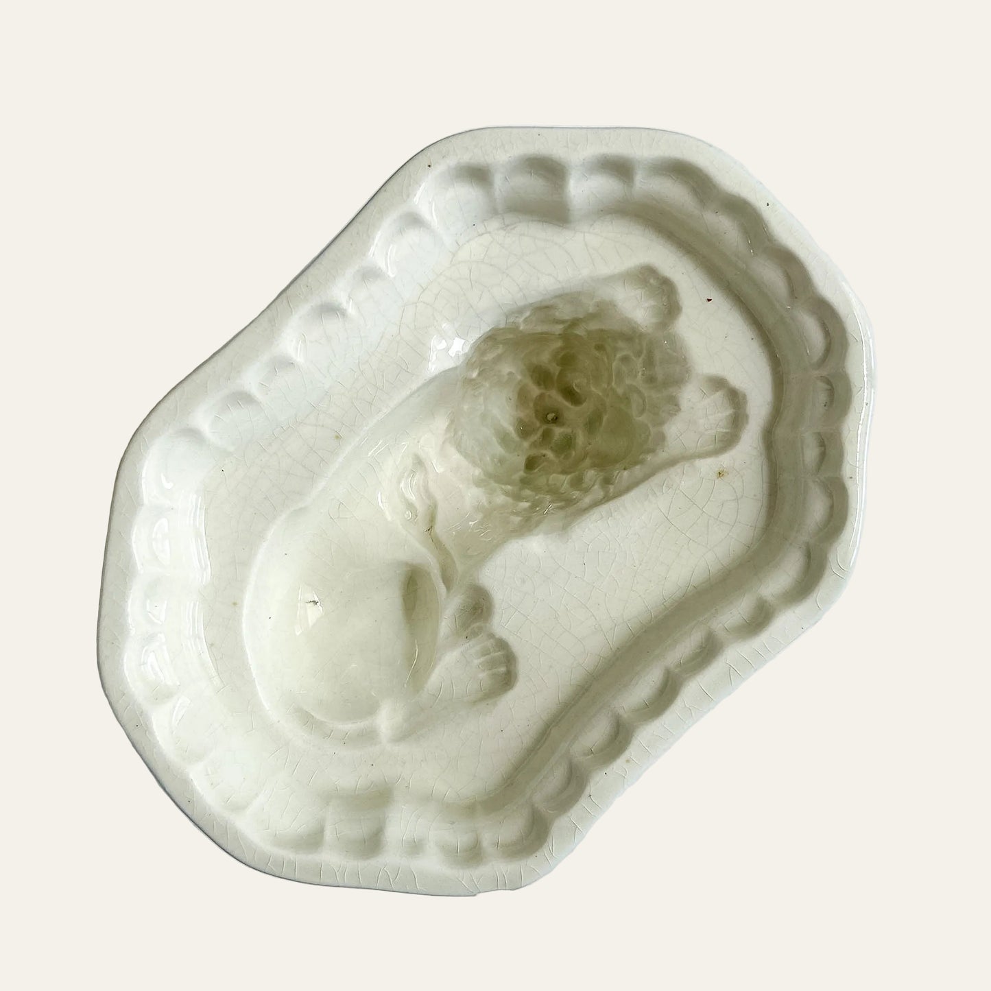 An antique ceramic dish with a raised relief of a sleeping figure, likely a child, in a pale cream glaze with visible crackle glaze patterns, set against a light textured background.