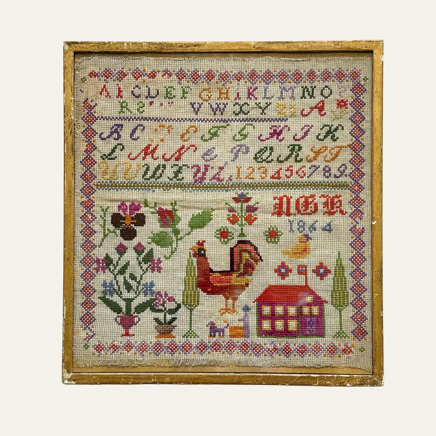 Framed cross-stitched sampler with alphabet, numbers, rooster, and house design on a textured white background.