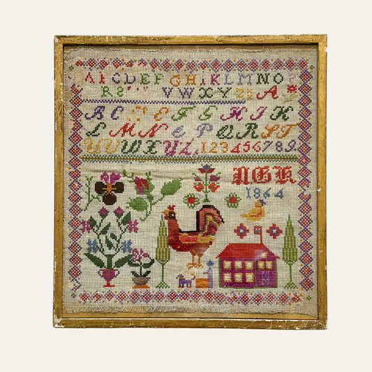Framed cross-stitched sampler with alphabet, numbers, rooster, and house design on a textured white background.