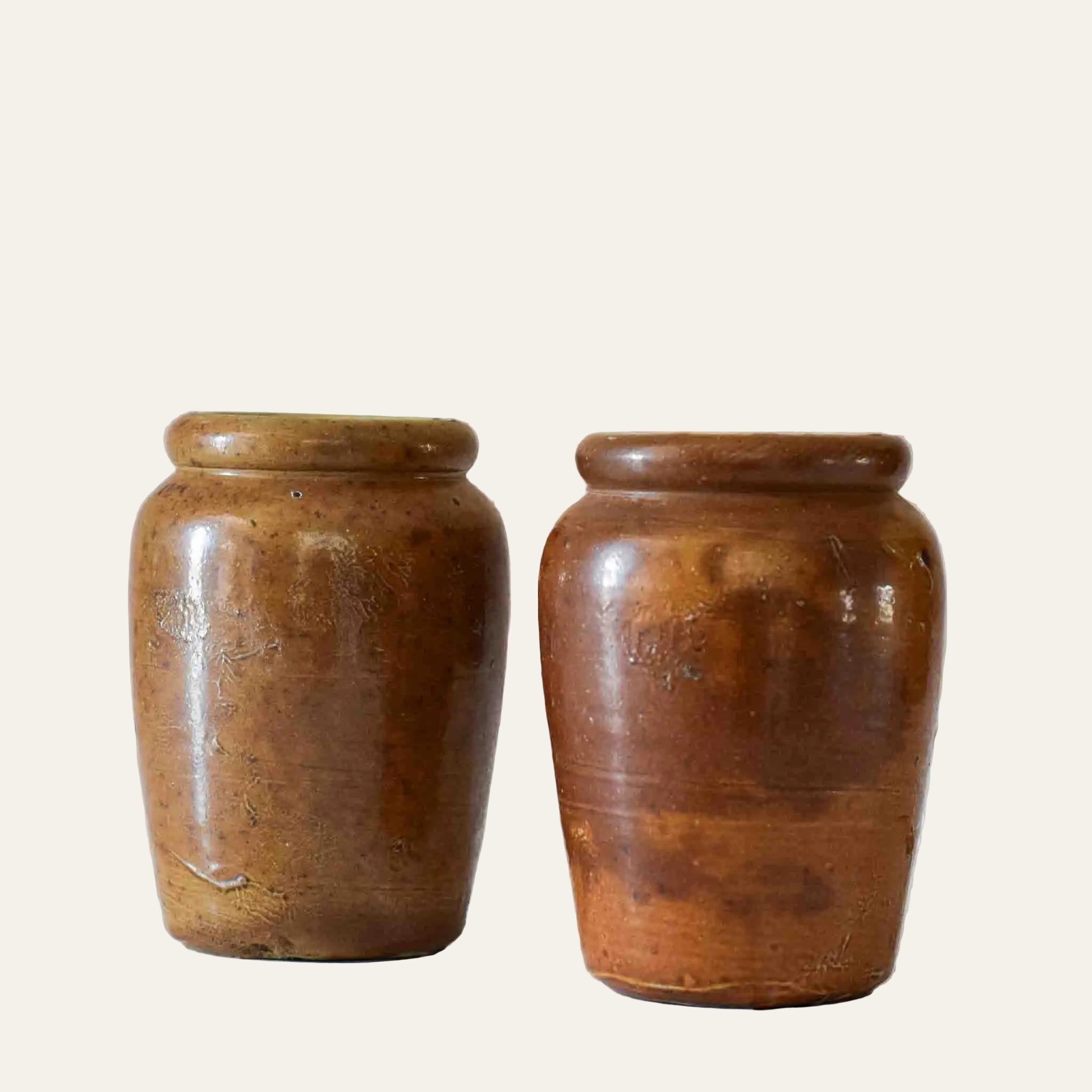 Two brown ceramic jars on a white tiled surface with a beige wall background