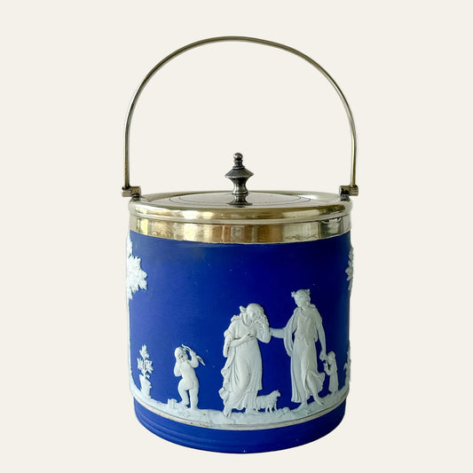 A classic Wedgwood-style ceramic tea caddy or storage jar in cobalt blue and white, featuring a detailed classical scene relief on the front, a silver metal lid, and a matching metal carrying handle.